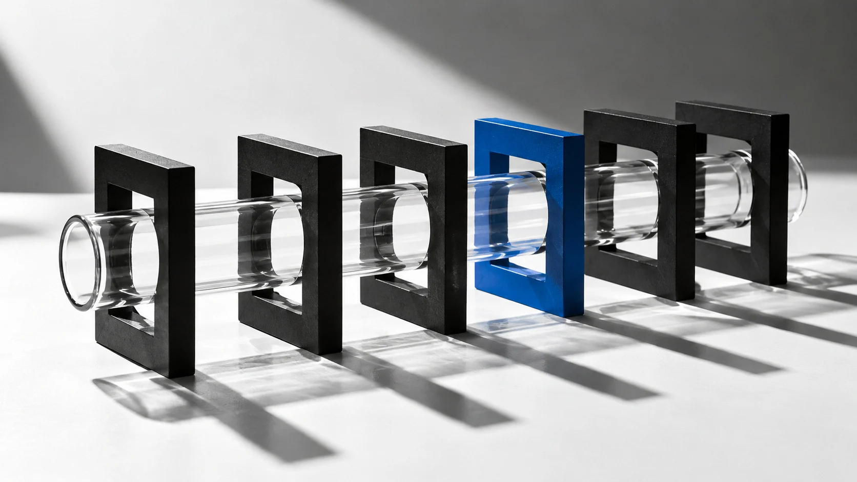 A glass tube is held in place by a series of black and one blue rectangular frames, casting shadows on a white surface.